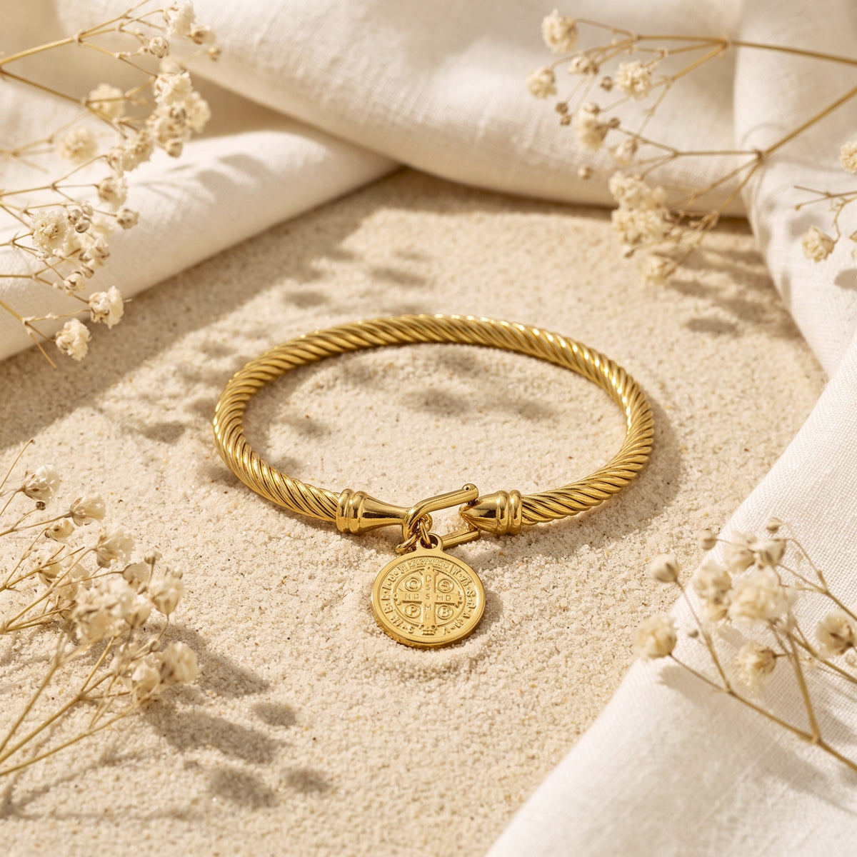 Angled view of the gold-tone twisted cable bracelet with its medal charm resting on sand beside white fabric.