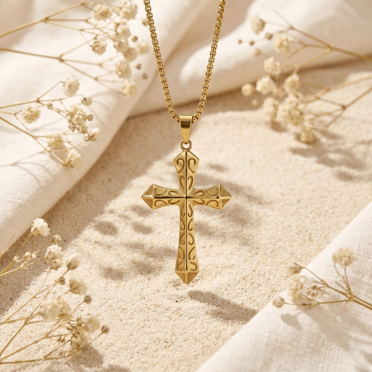 Front view of the gold-tone scrollwork cross pendant suspended vertically above sand with dried flowers.