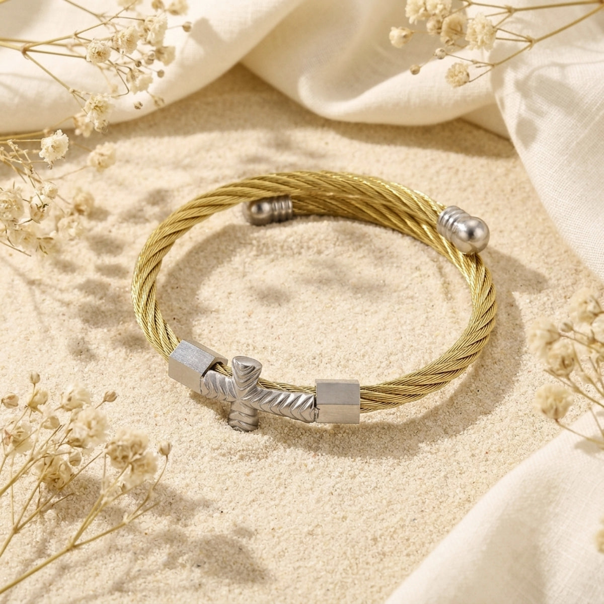 Overhead view of the gold-tone cable cuff with silver-tone cross and end caps on sand.