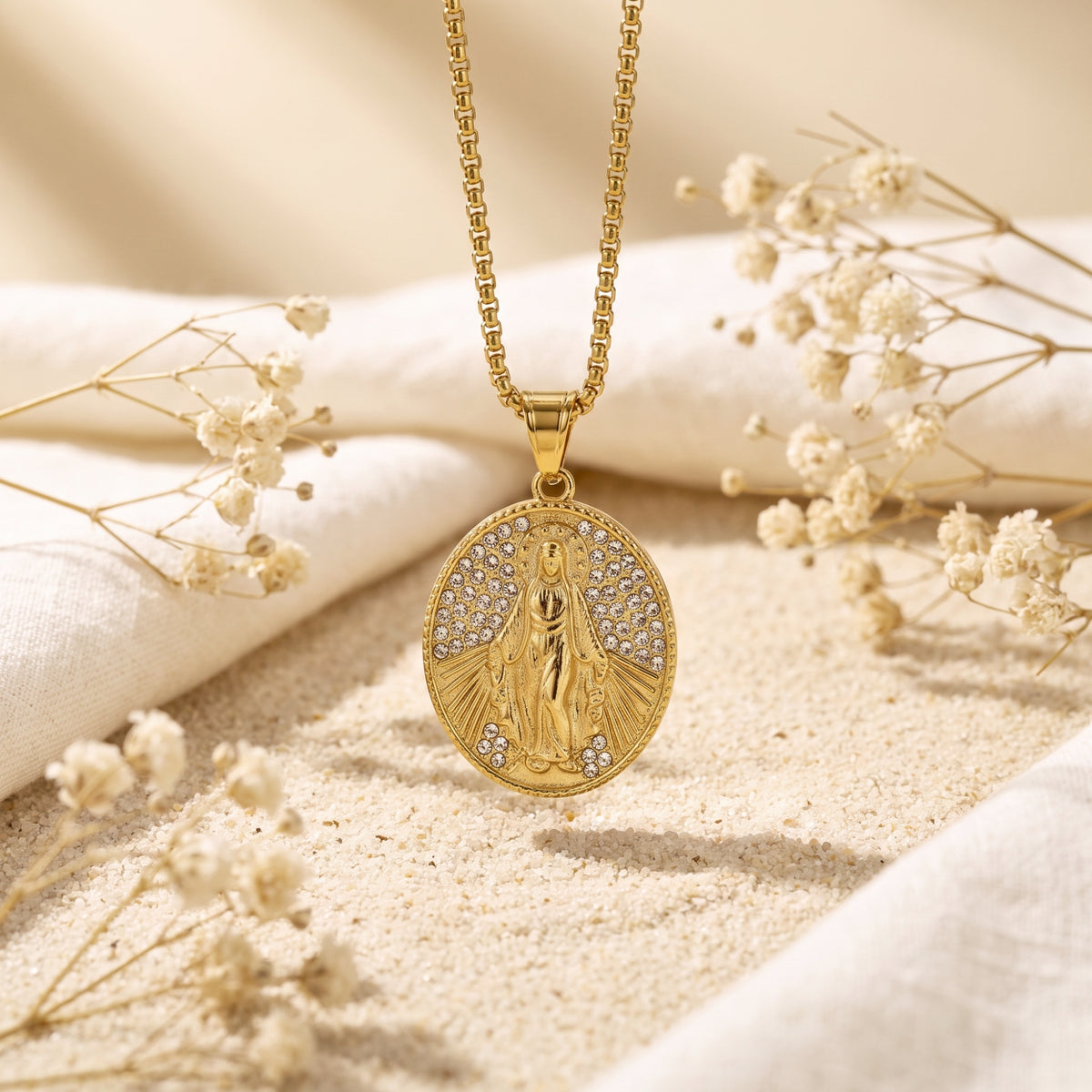 Front-facing gold-tone oval Miraculous Medal pendant hanging from a box chain over sand and white fabric.