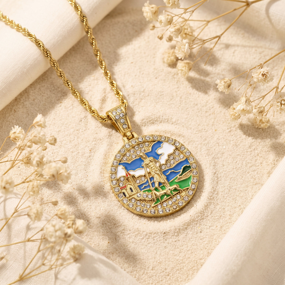 Gold-tone enamel medallion necklace resting diagonally on sand beside dried white flowers.