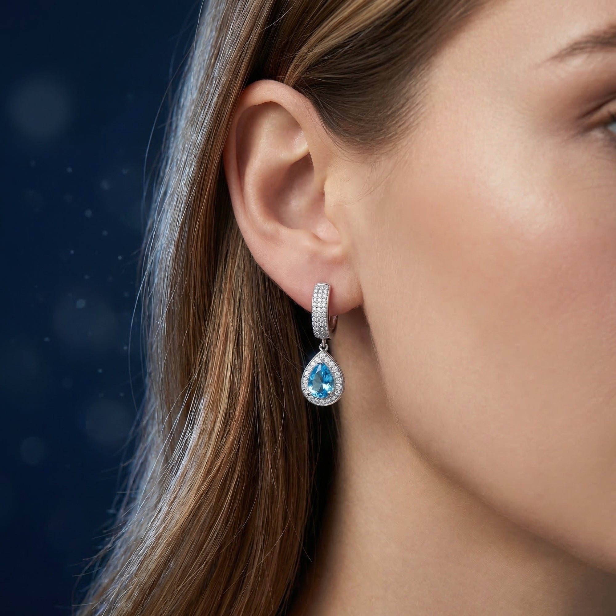 Profile view of a model wearing a silver pear cut Swiss blue topaz drop earring with a halo setting.