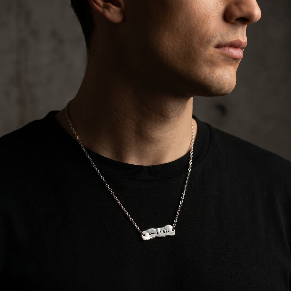 Wider view of a man wearing the Amor Fati bar necklace over a black T-shirt, shown in right-facing profile.