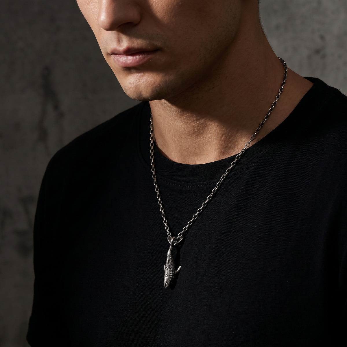 Three-quarter view of a man wearing the carved shark pendant necklace over a black T-shirt against a concrete wall.