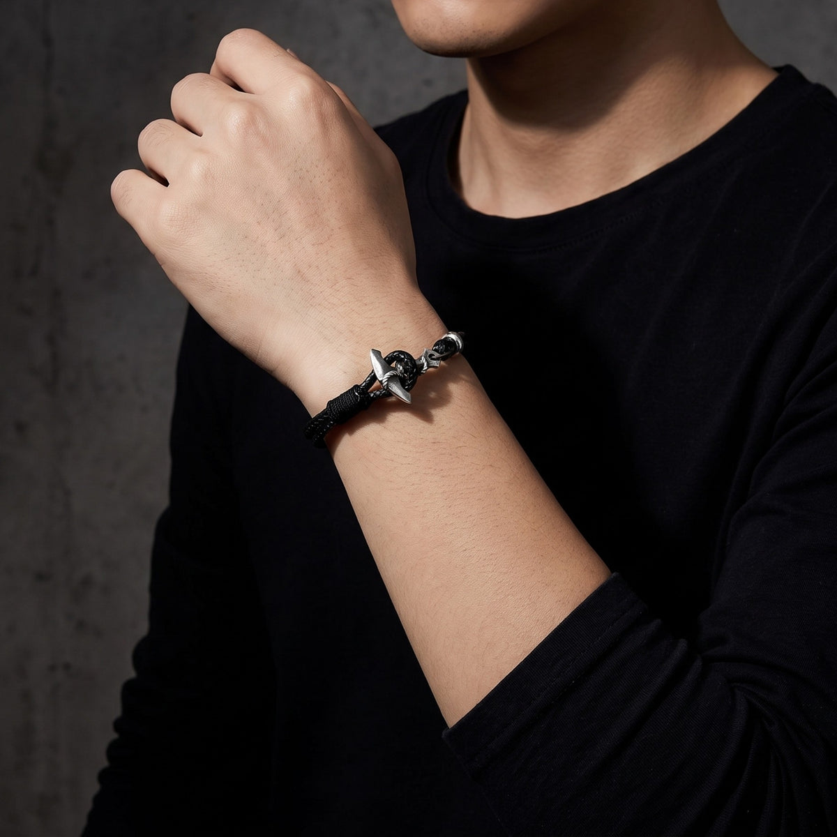 Person in a black long-sleeve shirt wearing a silver anchor bracelet against a dark wall.