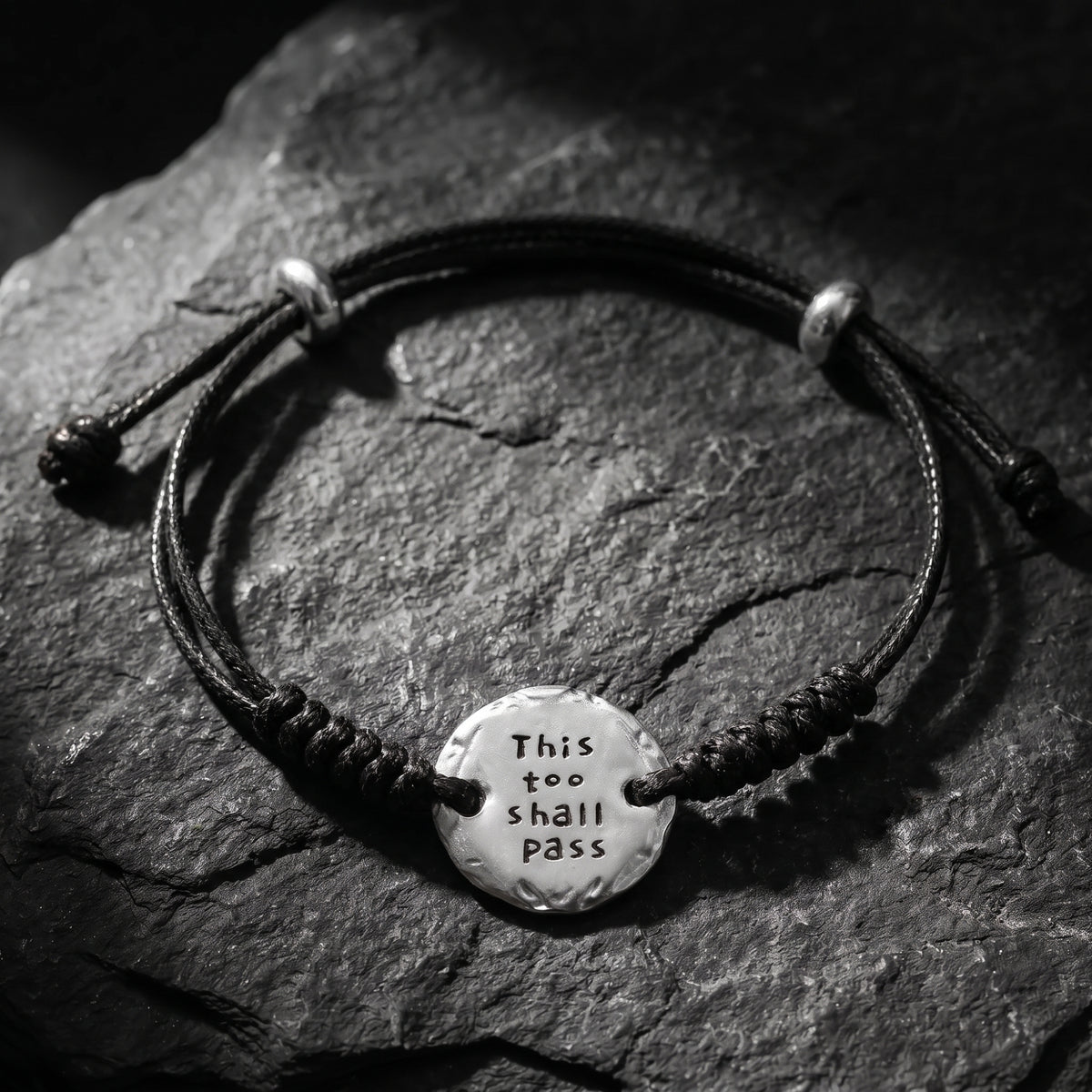 Black cord bracelet on dark stone with a silver medallion stamped “THIS TOO SHALL PASS.”