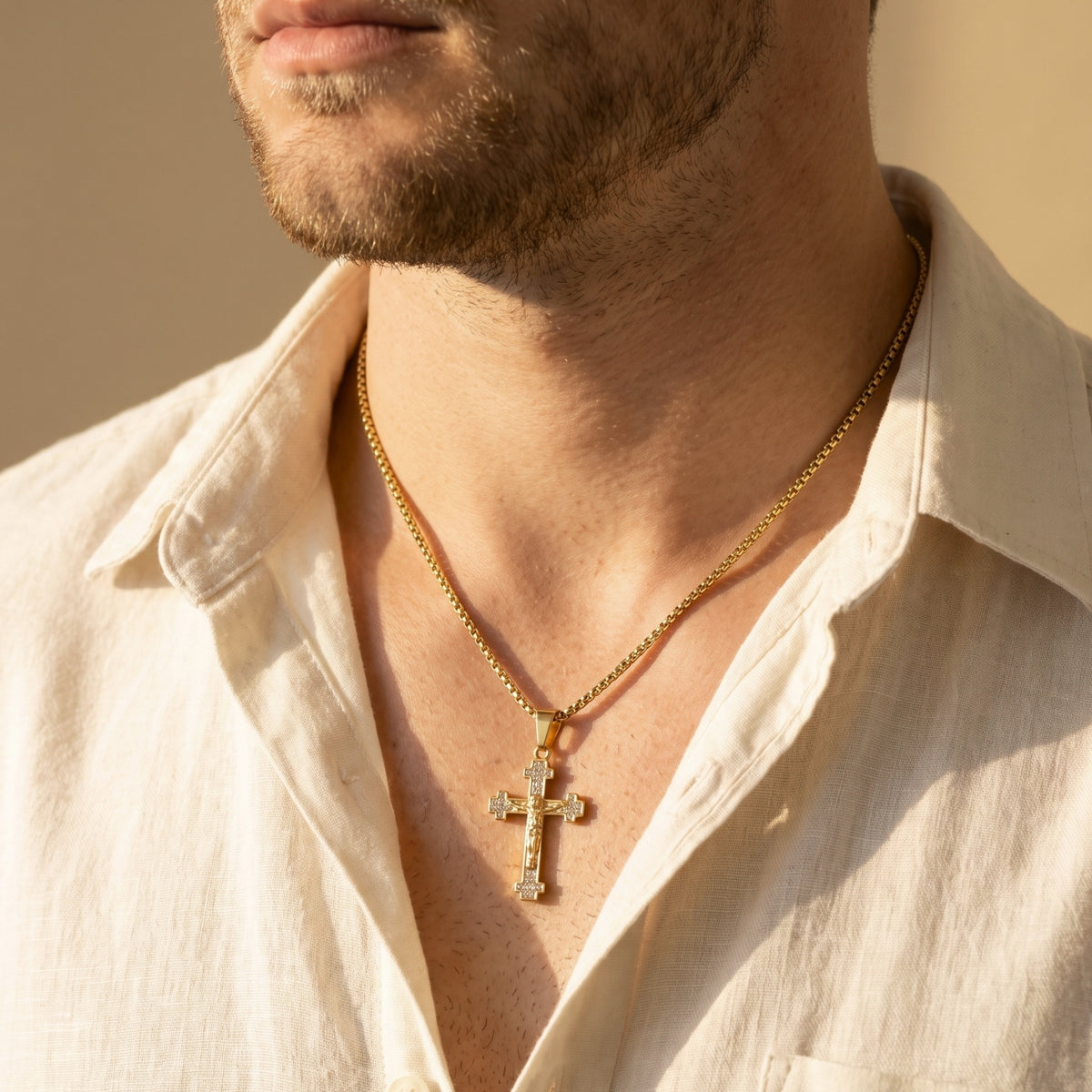 Close-up of a model wearing a gold crystal-accented crucifix necklace in warm sunlight with an open white shirt.