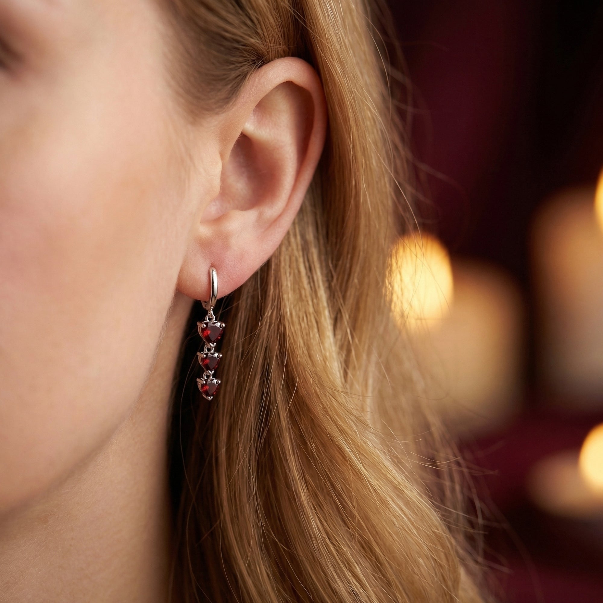 Side view of a model wearing silver heart drop earrings with three tiered red garnets, showing the jewelry's scale.