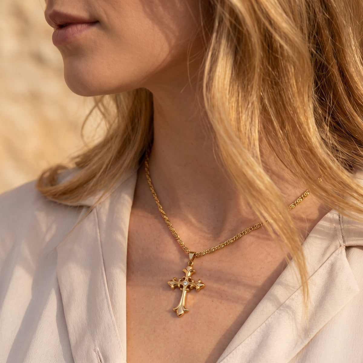 Model wearing a gold-tone filigree cross necklace over a light collared blouse in warm sunlight.