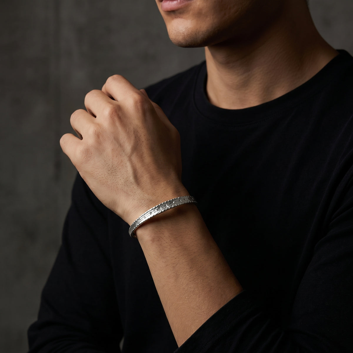 Etched silver cuff bracelet worn on a wrist as a hand rests across a black crew-neck shirt.