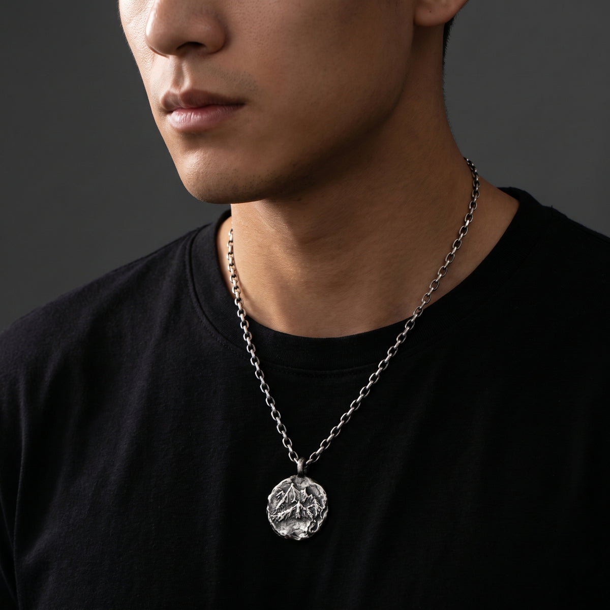 Three-quarter view of a model wearing a silver mountain-relief pendant over a black crewneck shirt.