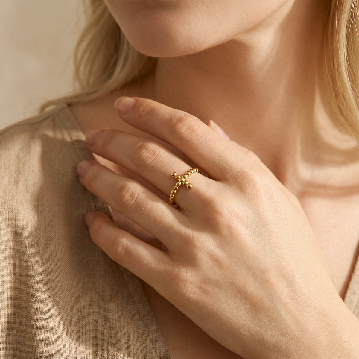 Gold-toned beaded cross ring worn on a finger as the model’s hand rests over a neutral linen garment.