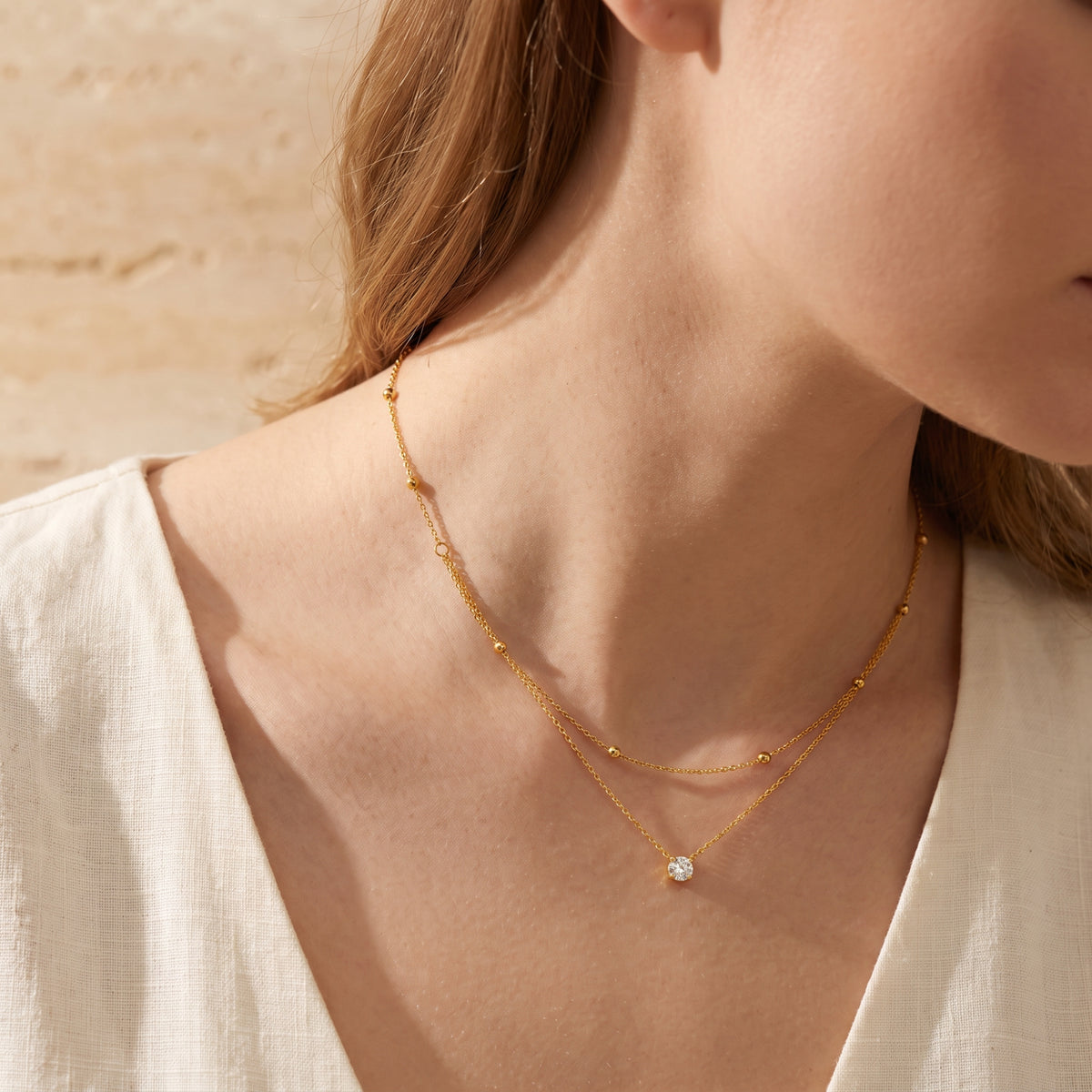 Three-quarter close view of a model wearing the gold-tone layered necklace with a beaded strand and clear solitaire pendant.