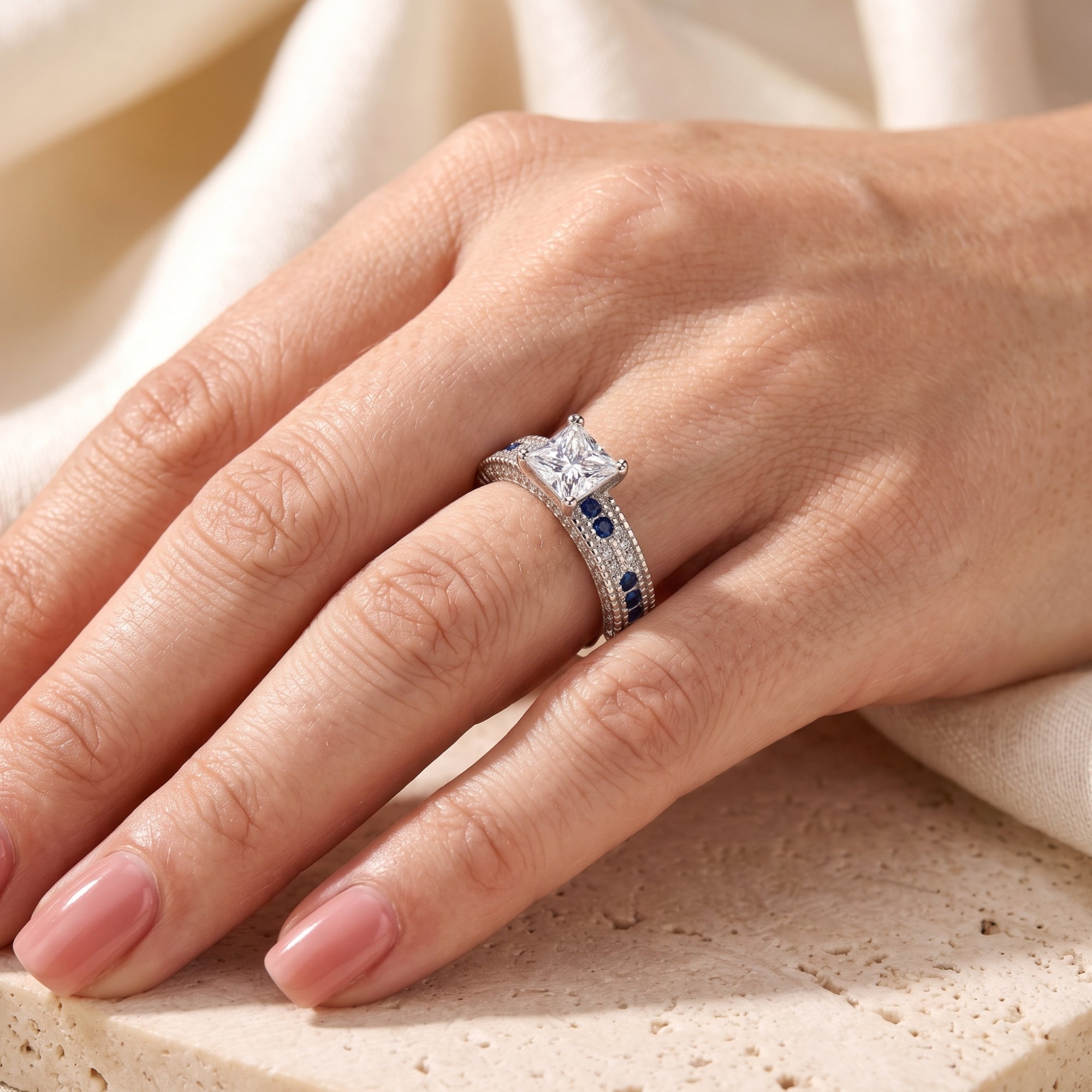 Silver princess-cut engagement ring with blue accents shown on a hand.