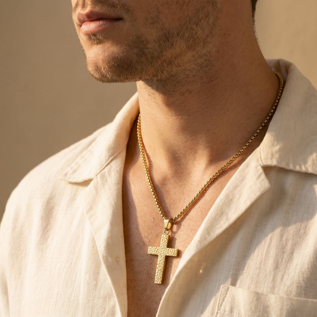 Man wearing a gold-tone hammered cross necklace over an open light-colored collared shirt.