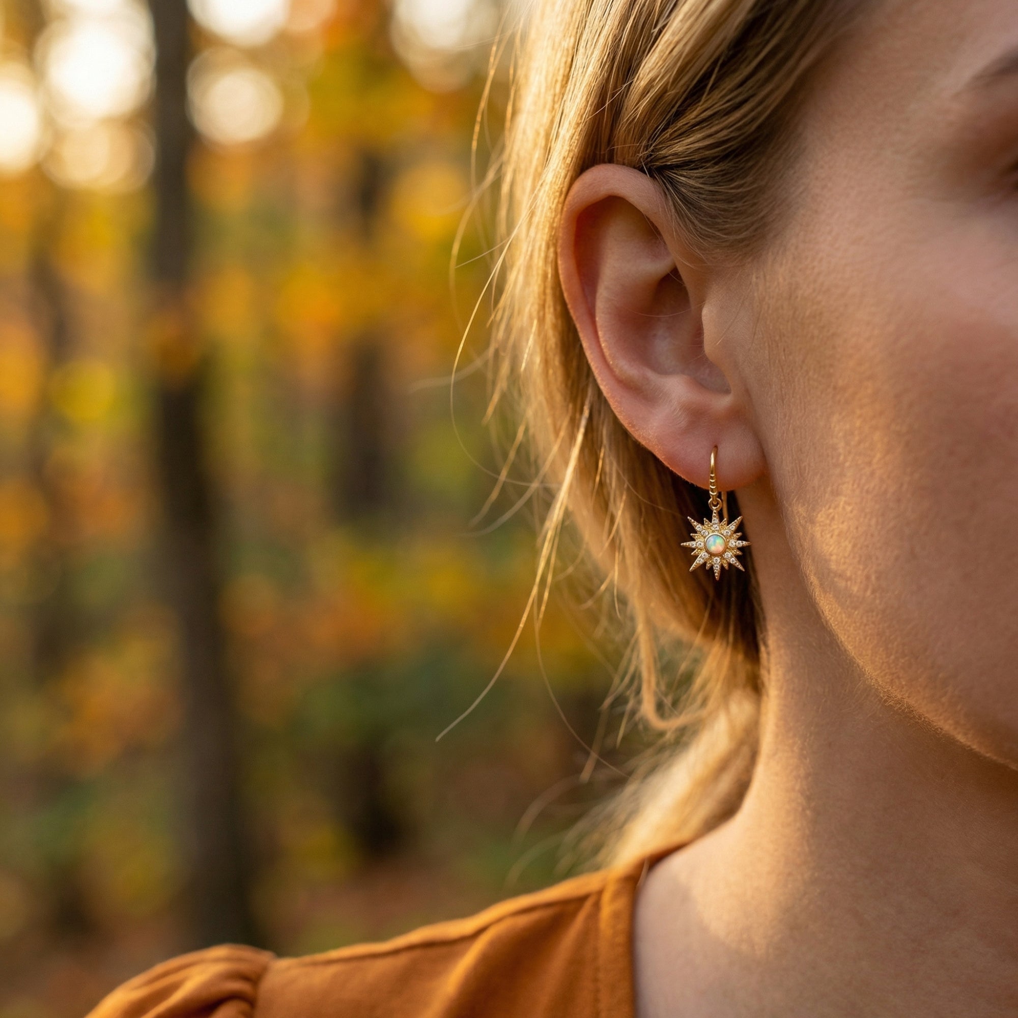 Model wearing a gold-tone starburst drop earring with a round white opal against a blurred autumn forest.