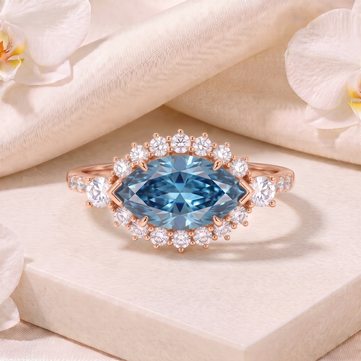 Marquise-cut blue moissanite halo ring in rose gold centered on a marble stand with silk draping.