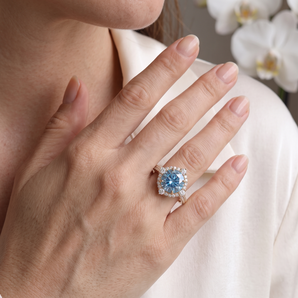 Close-up of a round blue moissanite halo engagement ring in rose gold worn on a model's hand.
