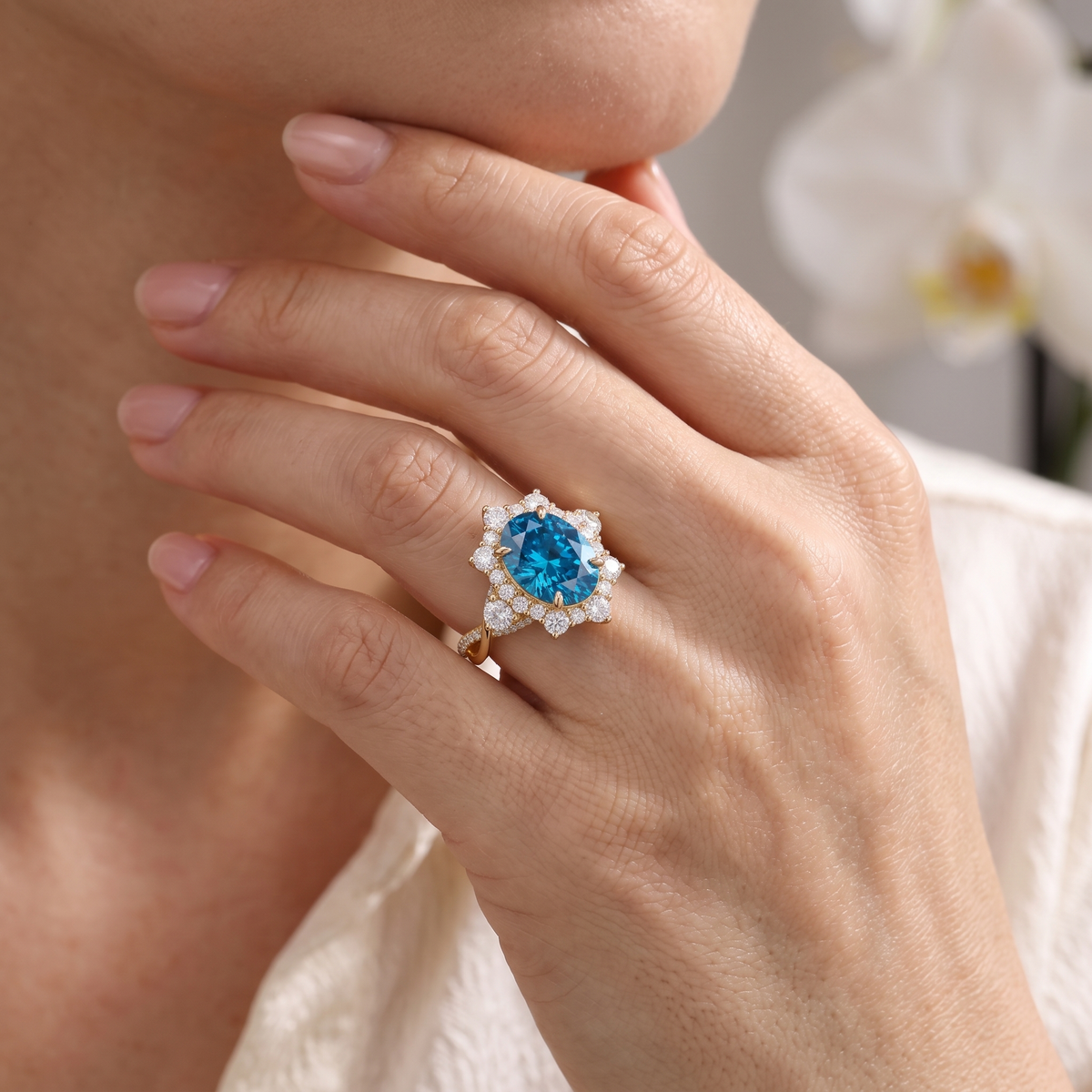 Close-up of a model's hand touching her chin wearing an oval blue moissanite halo ring in rose gold.