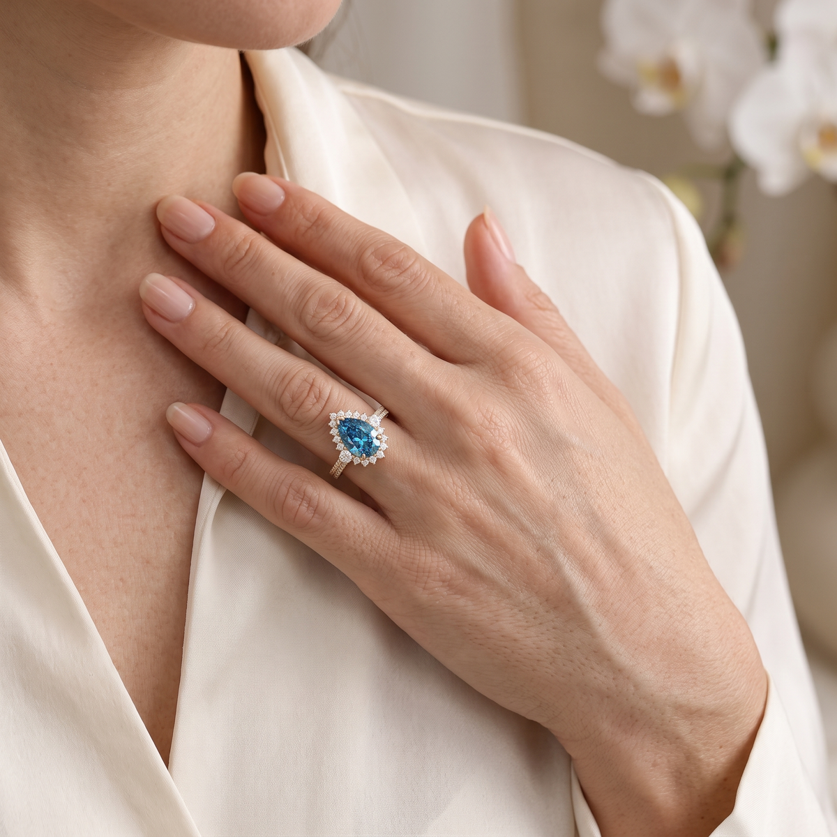 Model showcasing a pear cut blue moissanite halo engagement ring in rose gold against a cream silk blouse.