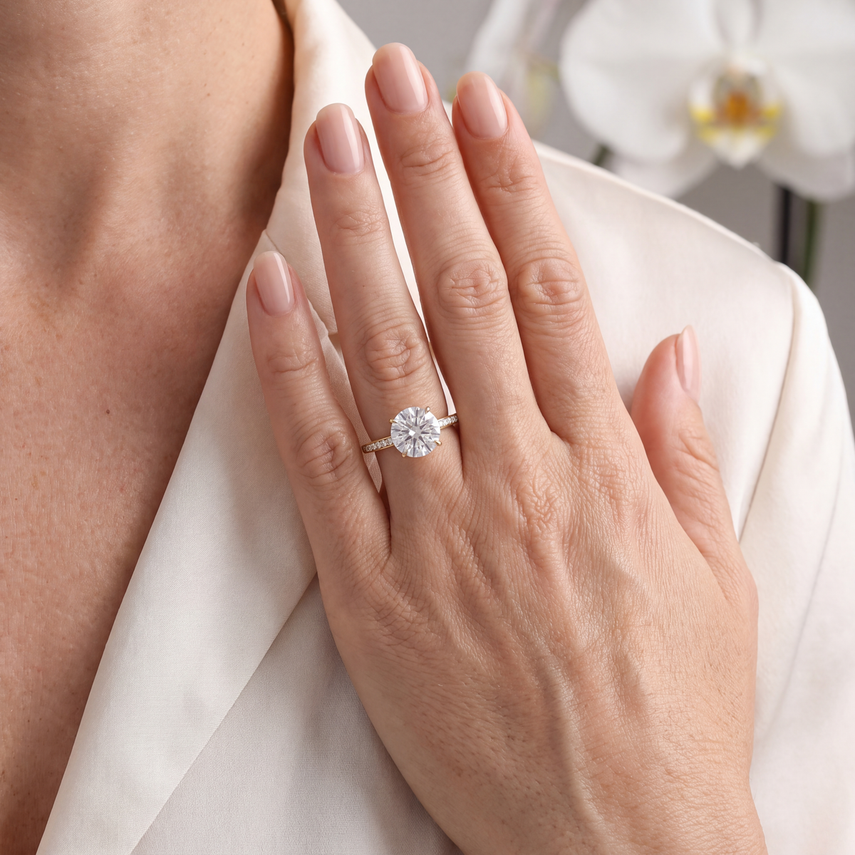 Yellow gold round moissanite ring with a pavé band displayed on a hand against a white blazer.