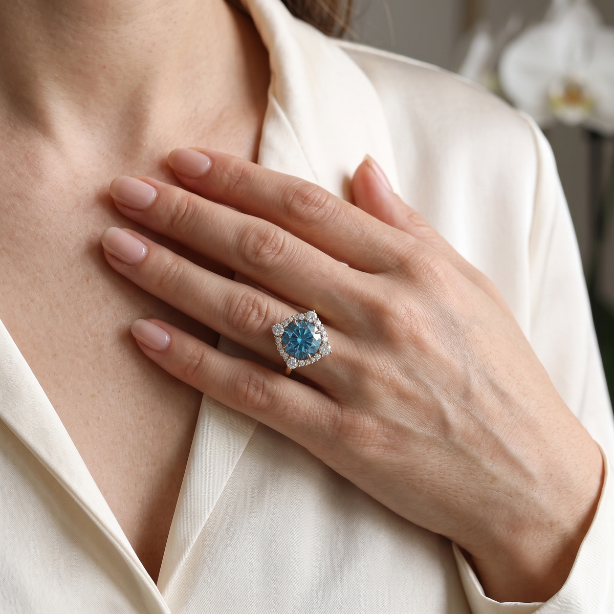 Woman wearing a rose gold halo ring with a blue moissanite center stone, hand placed on chest.