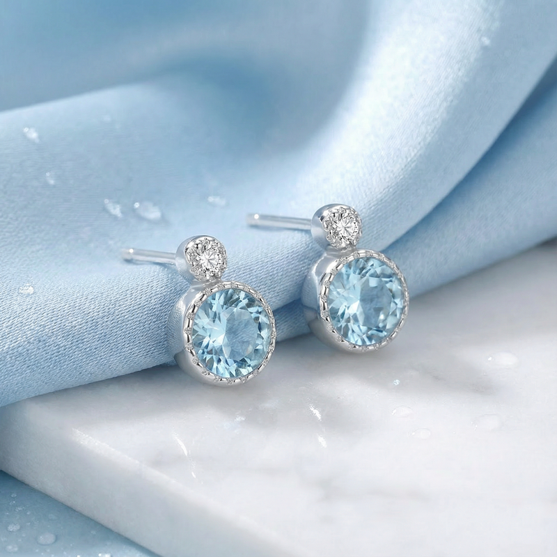 Front view of sterling silver simulated aquamarine studs with round accent stones on blue silk and marble.