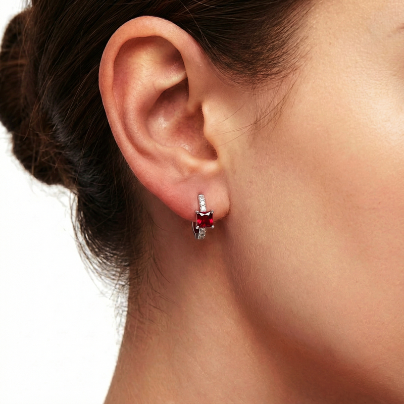 Close-up of a silver square huggie earring with a red simulated ruby worn on an ear.