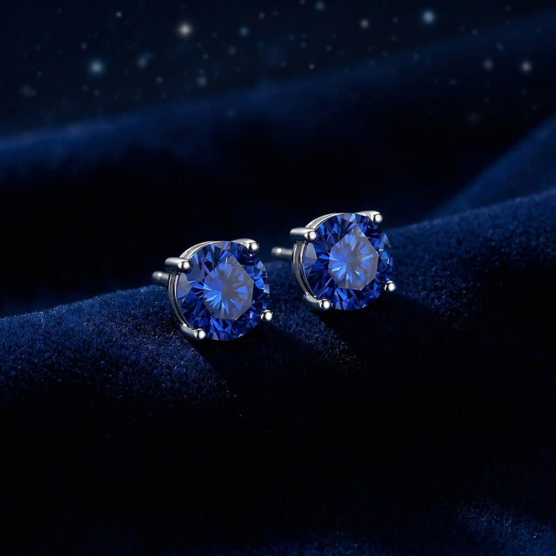 Side view of round blue sapphire stud earrings in silver settings resting on a textured blue velvet background.