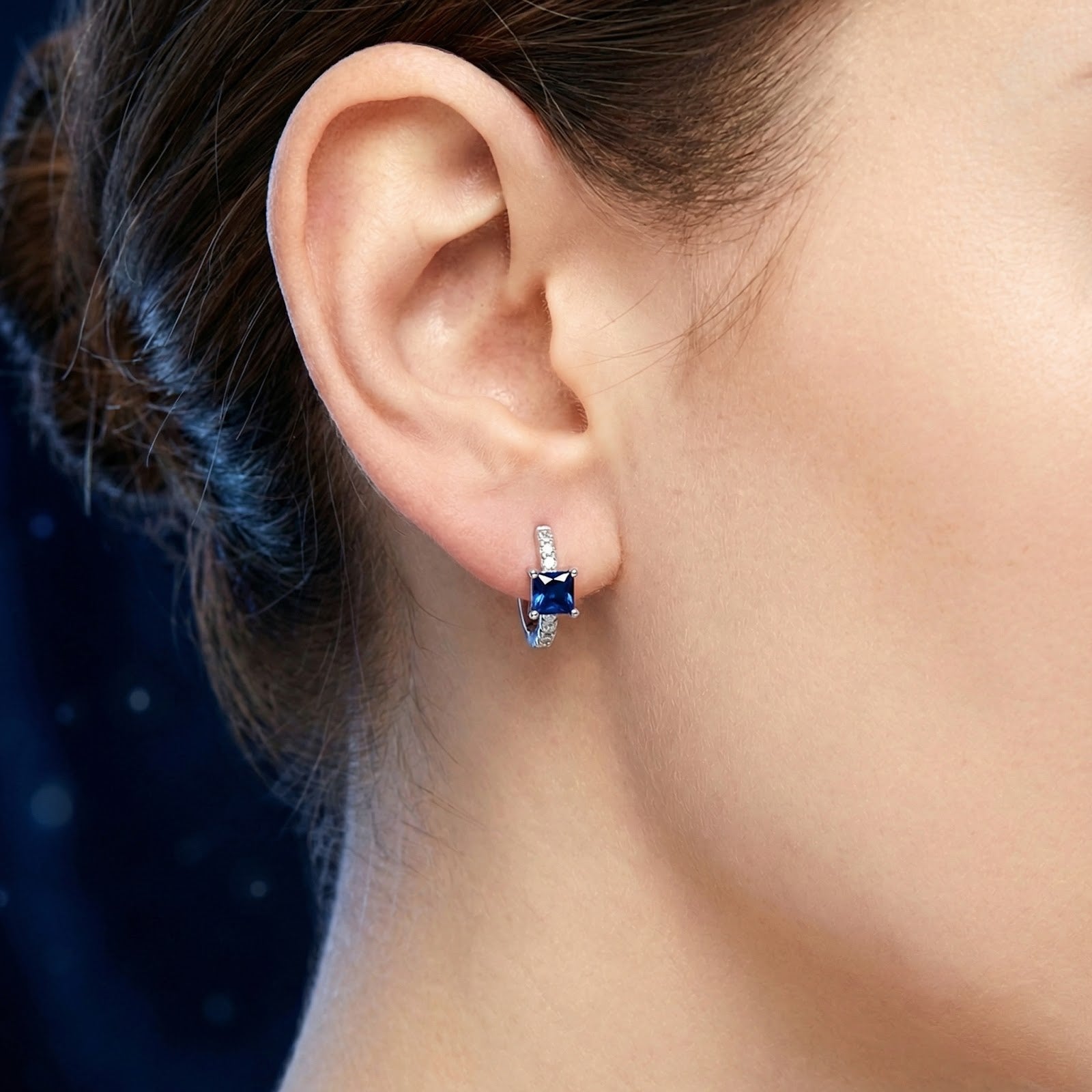 Close-up of a square-cut blue sapphire huggie earring in sterling silver worn on a woman's ear.