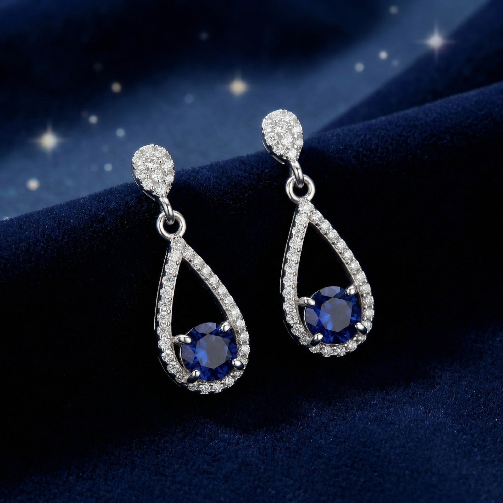 Sterling silver teardrop earrings with round simulated sapphires on a dark blue velvet background.