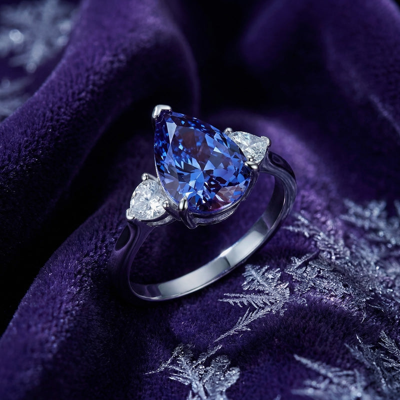 Angled view of a pear-cut simulated tanzanite three-stone ring in sterling silver on purple velvet.