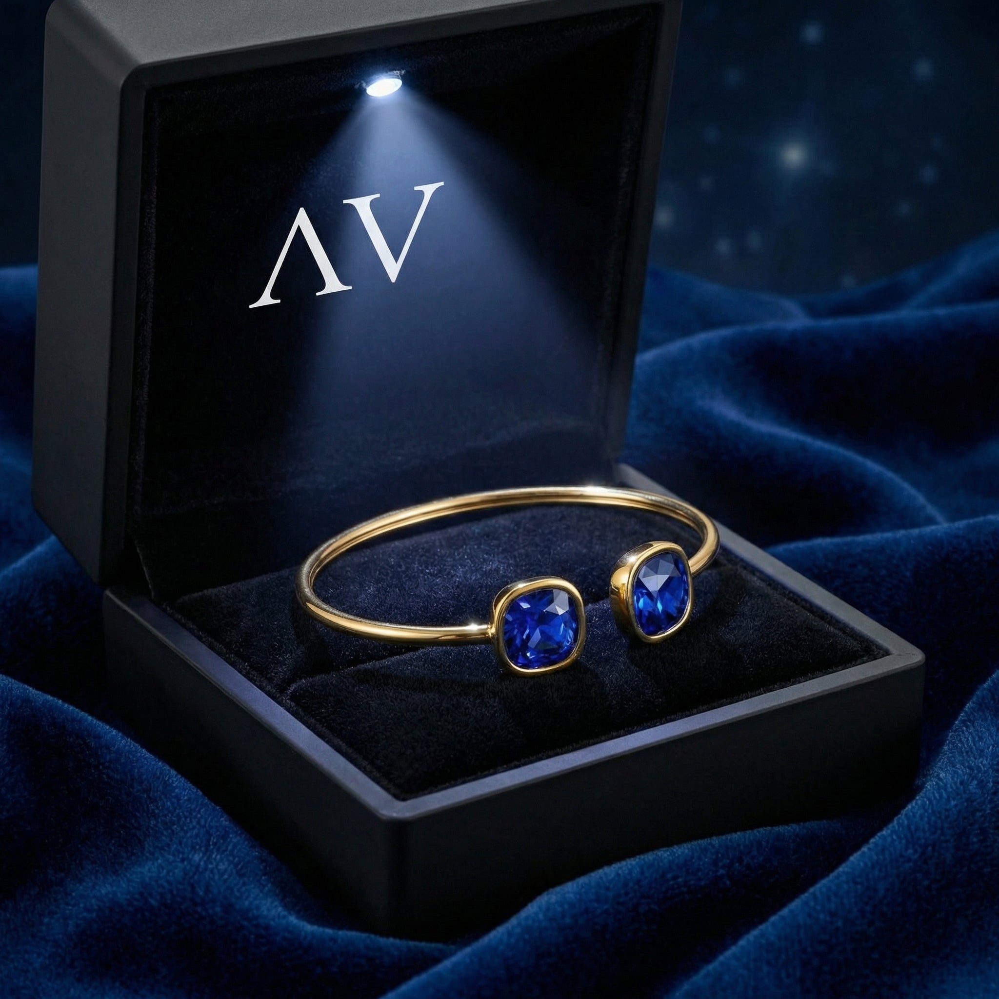 Gold-toned stainless steel cuff with cushion-cut blue sapphires in a lighted black jewelry box.