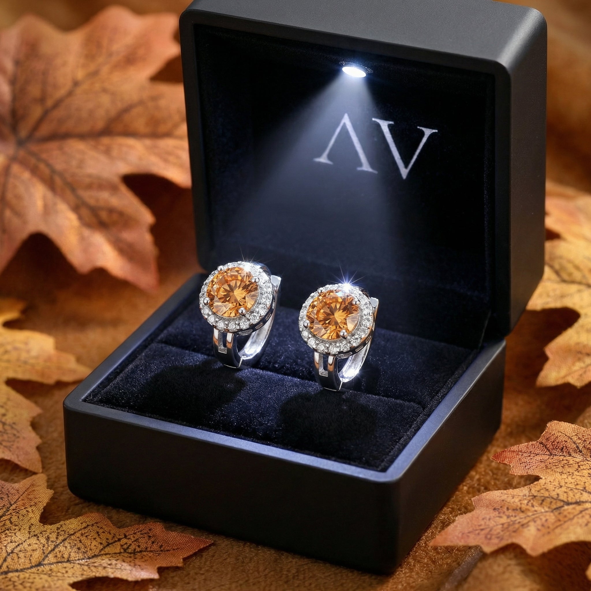 Honey moissanite halo earrings in sterling silver displayed in a lighted black jewelry box with leaves.