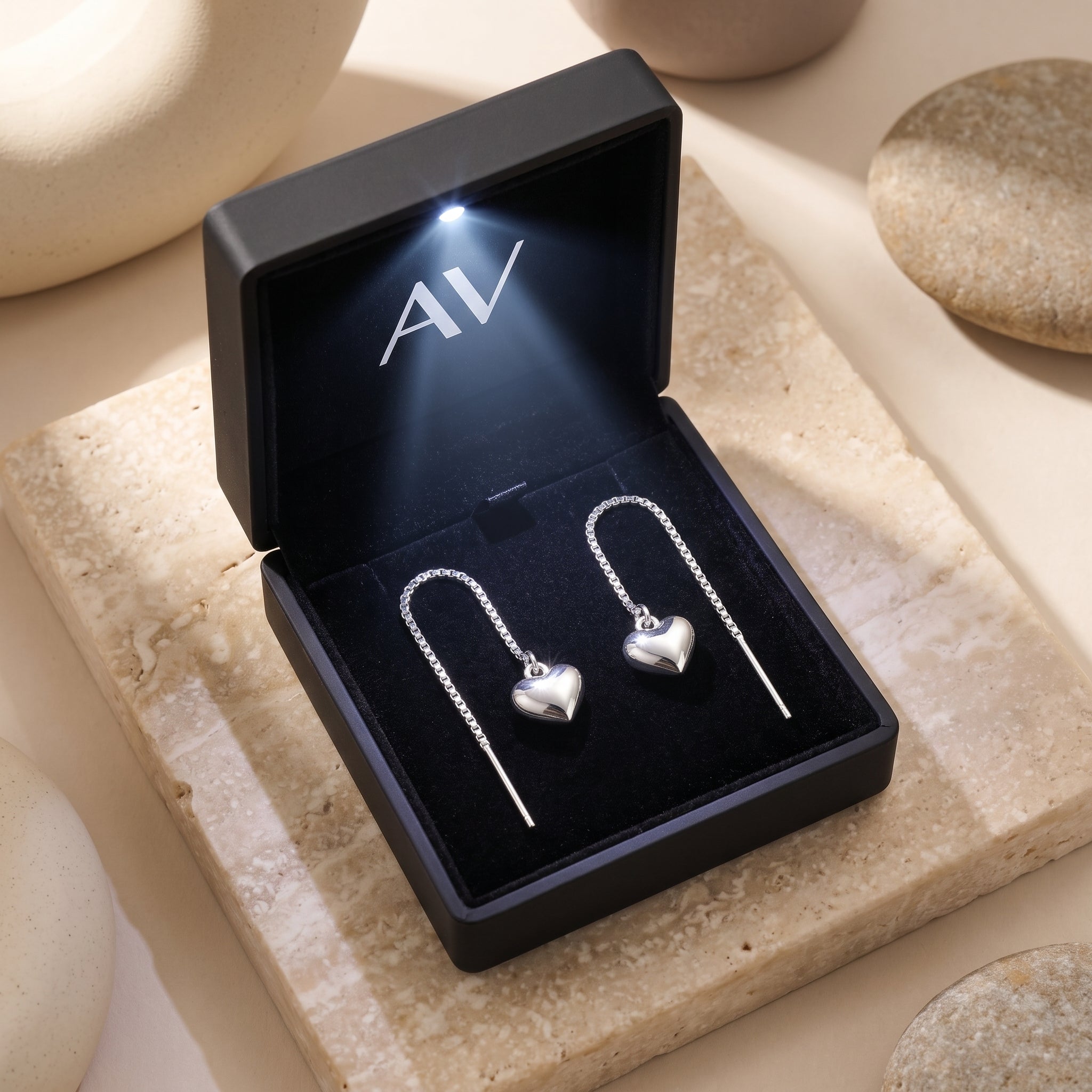Silver heart threader earrings presented in a black LED-lit jewelry box on a stone background.
