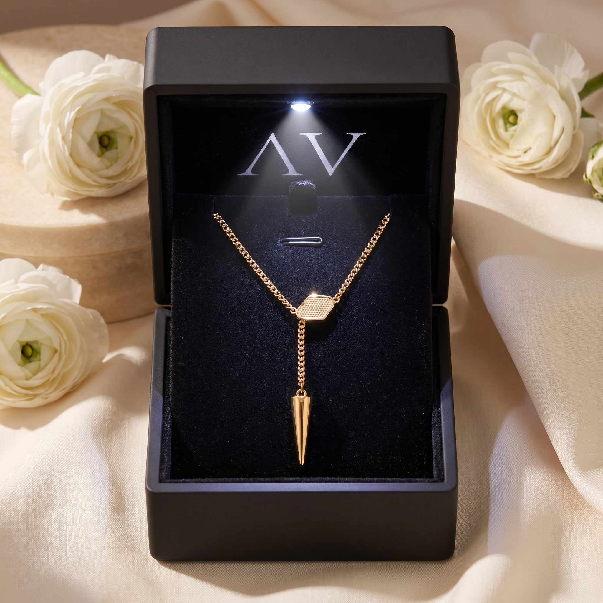 Gold lariat necklace in a black LED-lit jewelry box with white floral background.