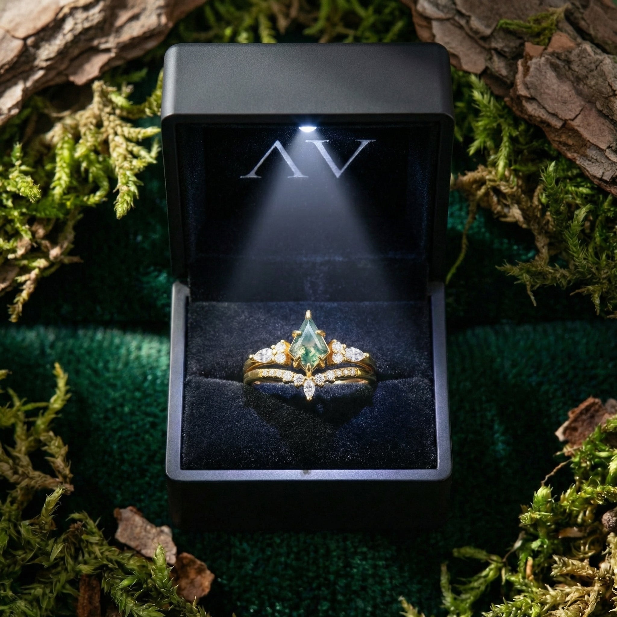 Kite-cut moss agate ring set displayed in a lighted black jewelry box surrounded by natural moss and bark.