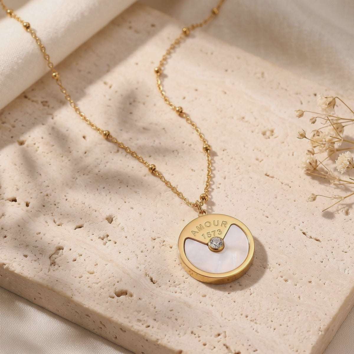 Close-up of the gold-tone L'Amour medallion necklace with mother-of-pearl inlay on a textured beige stone surface.