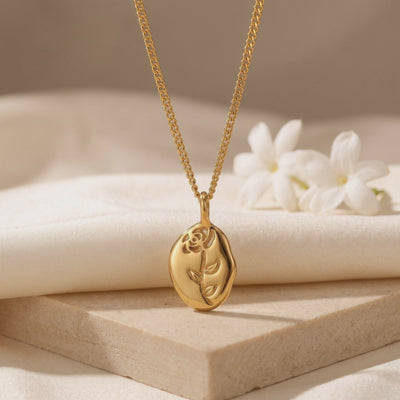 Gold-tone necklace with a curb chain and rose-etched oval pendant displayed on a beige stone slab beside white flowers.
