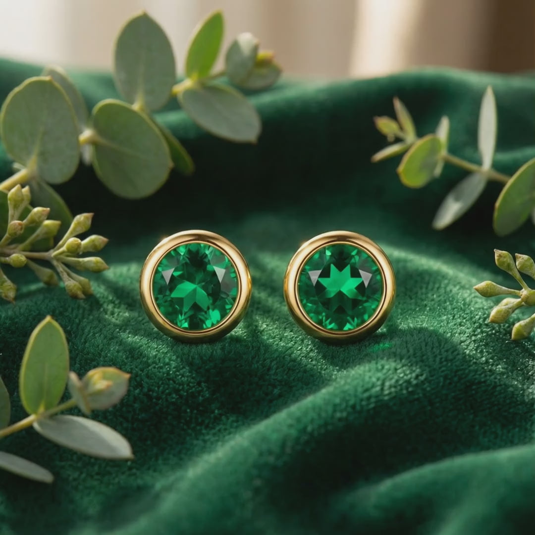 Pair of round green simulated emerald stud earrings in solid gold on green velvet, framed by leafy foliage.