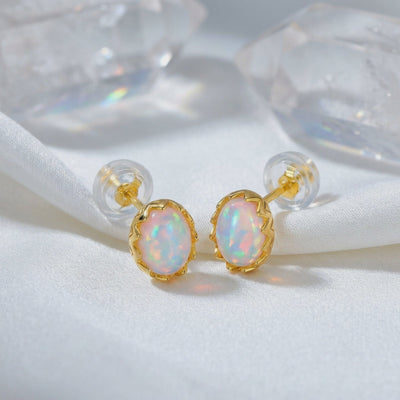 Pair of oval white opal stud earrings in solid gold with clear silicone backs on white fabric.