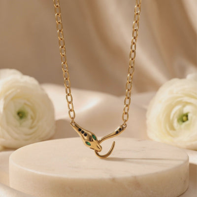 Yellow gold-tone link necklace with a green-accented snake clasp displayed above a light-colored pedestal and white flowers.