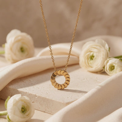 Gold-tone overlapping-disk pendant necklace displayed against white fabric, a stone block, and white ranunculus blooms.