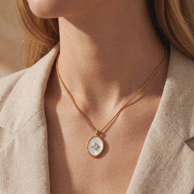 Gold-tone necklace with an oval mother-of-pearl pendant and square center stone, worn over a beige blazer.