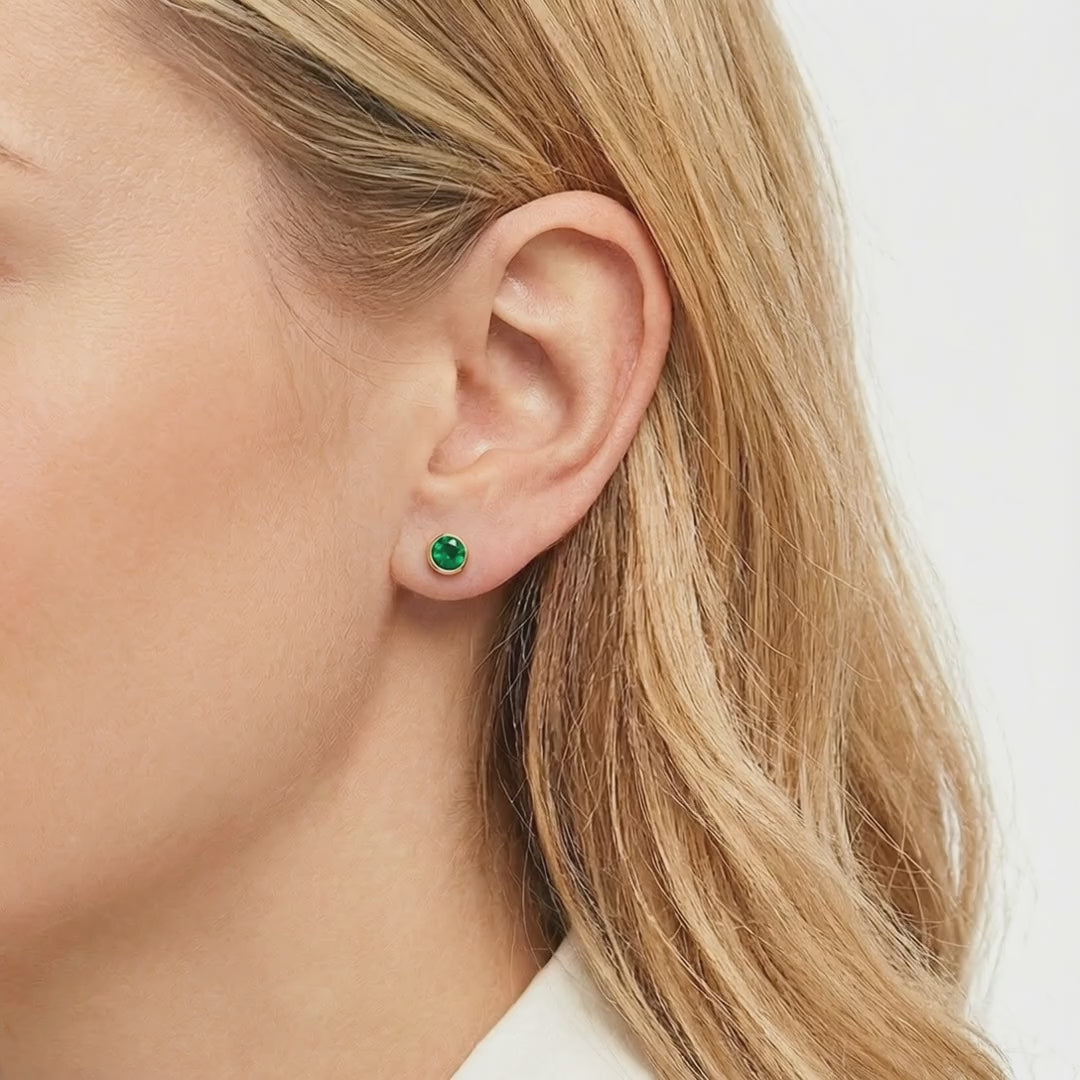 Close-up side view of a woman wearing one round green simulated emerald stud earring in a solid gold bezel setting.
