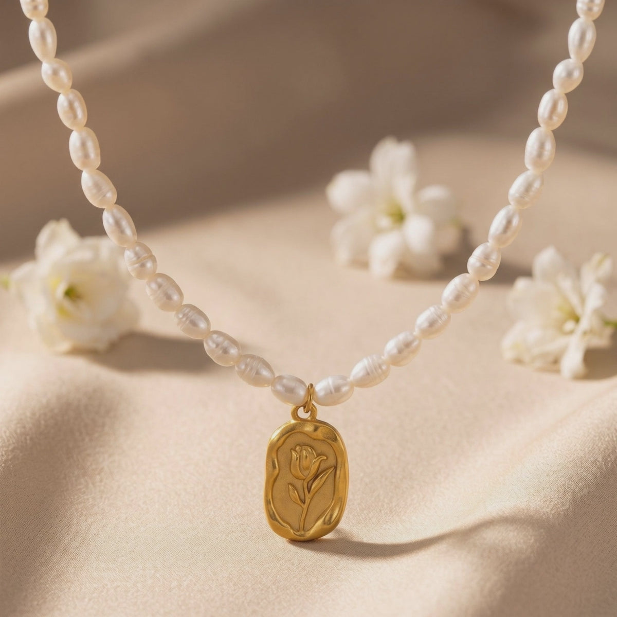 Pearl necklace arranged on soft fabric with a gold-toned rose pendant and white flowers nearby.