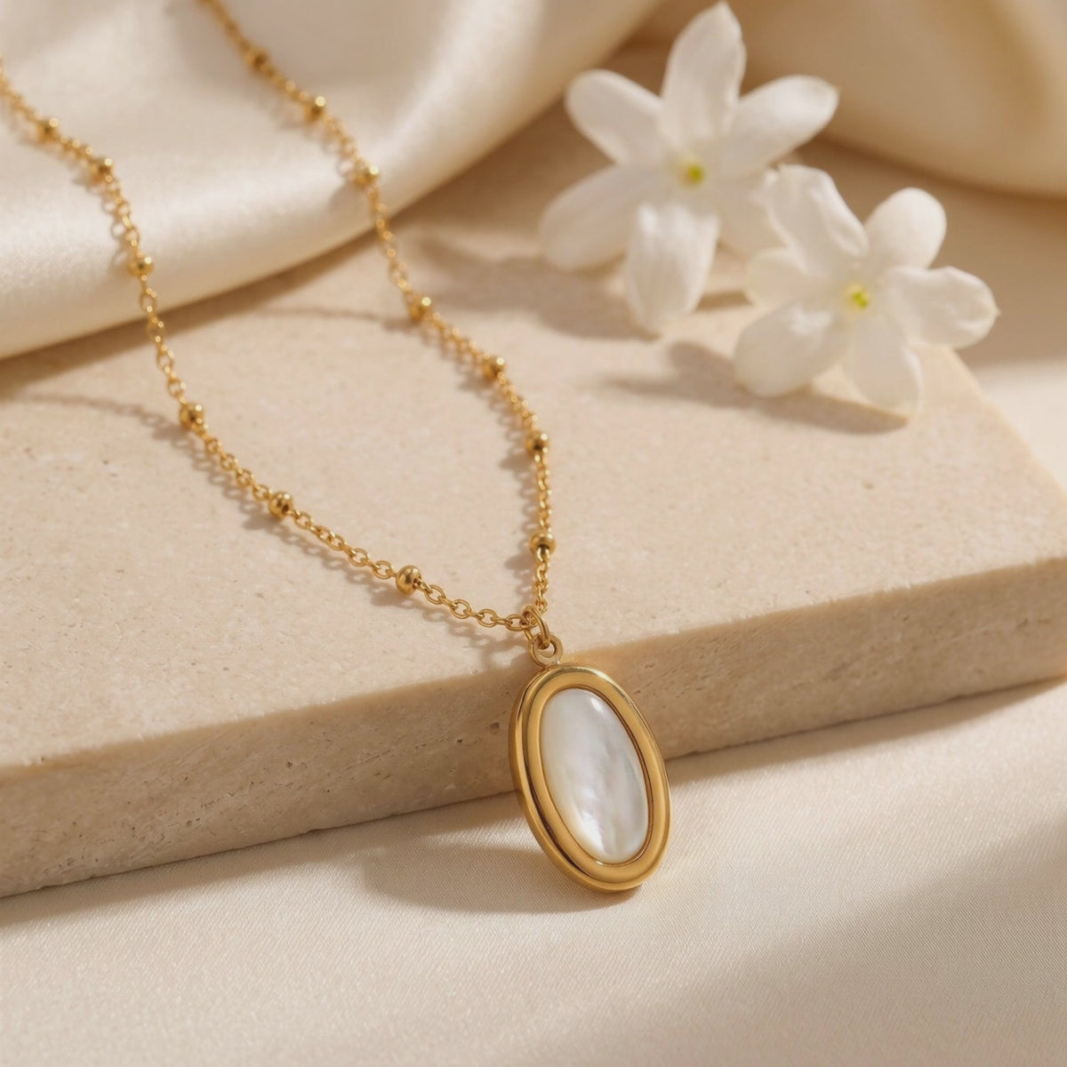 Gold mother-of-pearl oval pendant necklace displayed on a beige stone slab with white flowers.
