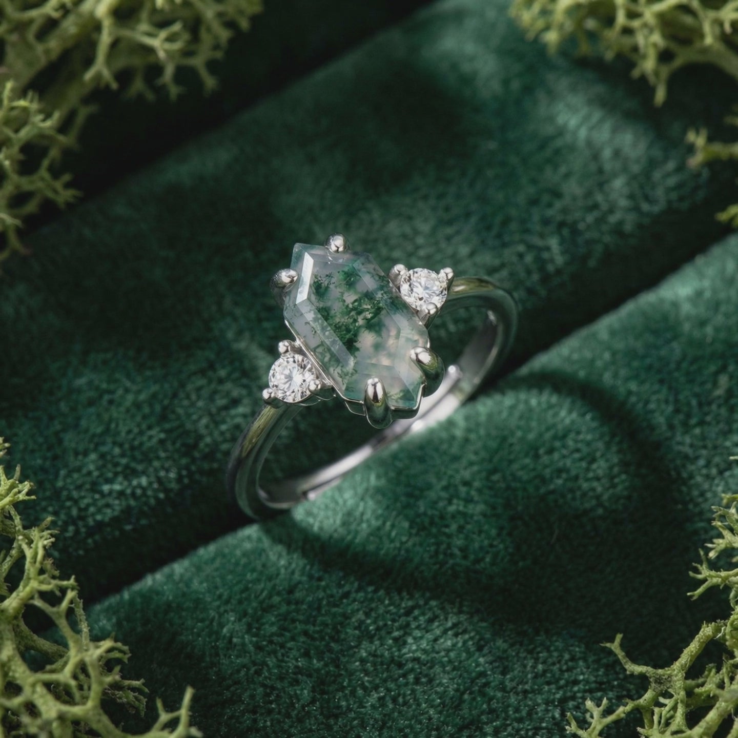 Solid white gold ring with elongated hexagonal moss agate center and two round side stones on dark green velvet.