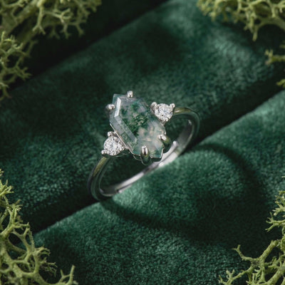 Solid white gold ring with elongated hexagonal moss agate center and two round side stones on dark green velvet.