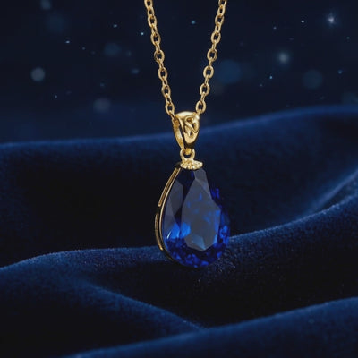 Close-up of a faceted pear-shaped simulated sapphire pendant necklace in solid gold on dark blue velvet.